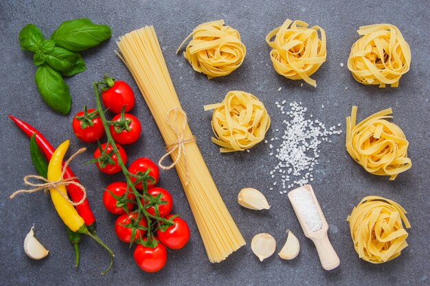 Chili peppers, a bunch of tomatoes, salt, black pepper, garlic, leaves and spaghetti and tagliatelle pasta on a gray surface. top view.