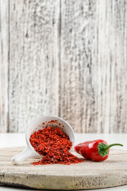 Chili powder with red pepper and wooden piece in a white scoop on white and wooden grunge wall, side view.