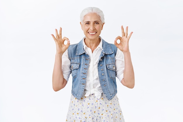 회색 머리를 한 걱정 없는 행복한 노부인은 모든 것이 괜찮다는 것을 보여주는 데님 조끼 드레스를 입고 웃고 있습니다.