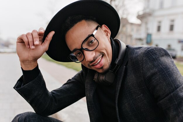 Chilling african young man in glasses posing with confident face expression. Outdoor shot of relaxing guy in hat and woolen suit enjoying photoshoot.