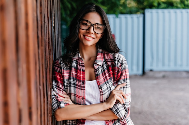 Chilling latin girl posing on the street with arms crossed and laughing. Beautiful brunette woman standing with shy smile.