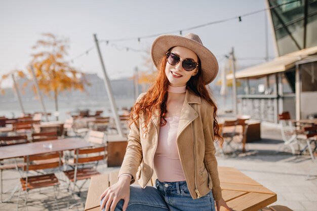 Chilling young woman in casual outfit looking through sunglasses with smile sitting on table