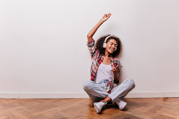 Chilling young woman sitting on the floor and listening music. Amazing african girl posing with legs crossed while enjoying favorite song.