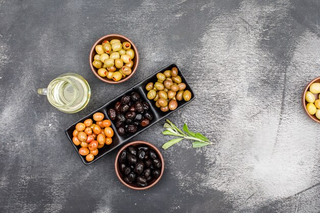 Chilly olives assortment in a black plate and clay bowls with a jar of olive oil and olive leaves top view on dark grey grunge