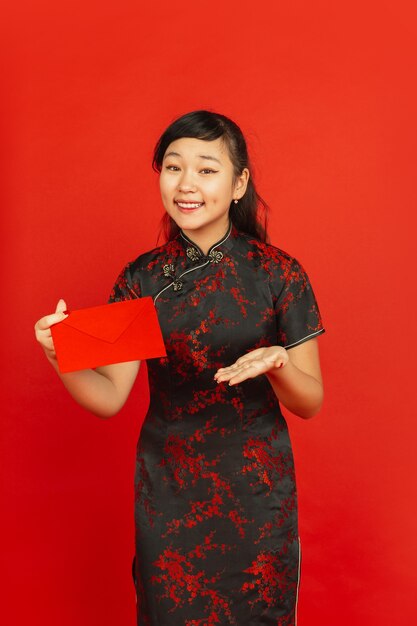 Chinese New Year 2020. Asian young girl's portrait isolated on red background. Female model in traditional clothes looks happy, smiling and showing red envelope. Celebration, holiday, emotions.