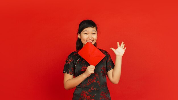 Chinese New Year 2020. Asian young girl's portrait isolated on red background. Female model in traditional clothes looks happy, smiling and showing red envelope. Celebration, holiday, emotions.