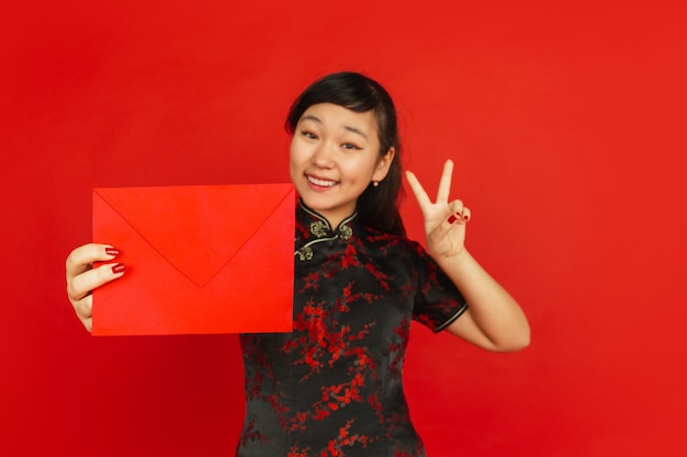 Chinese New Year 2020. Asian young girl's portrait isolated on red background. Female model in traditional clothes looks happy, smiling and showing red envelope. Celebration, holiday, emotions.