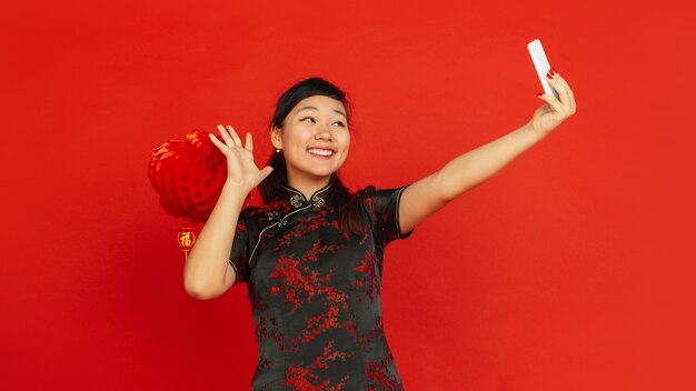 Chinese New Year 2020. Asian young girl's portrait isolated on red background. Female model in traditional clothes looks happy and taking selfie with decoration. Celebration, holiday, emotions. Flyer.