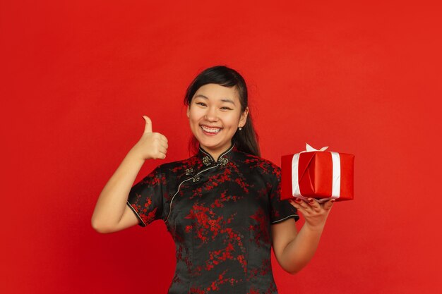 Chinese New Year 2020. Asian young girl's portrait isolated on red background. Female model in traditional clothes looks happy with giftbox. Celebration, holiday, emotions. Showing nice, smiling.