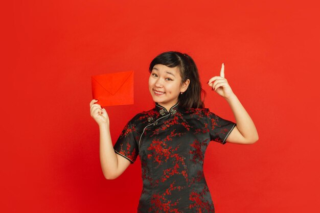 中国の旧正月。赤い背景で隔離のアジアの若い女の子の肖像画。伝統的な服を着た女性モデルは、幸せそうに見え、笑顔で赤い封筒を指しています。お祝い、休日、感情。