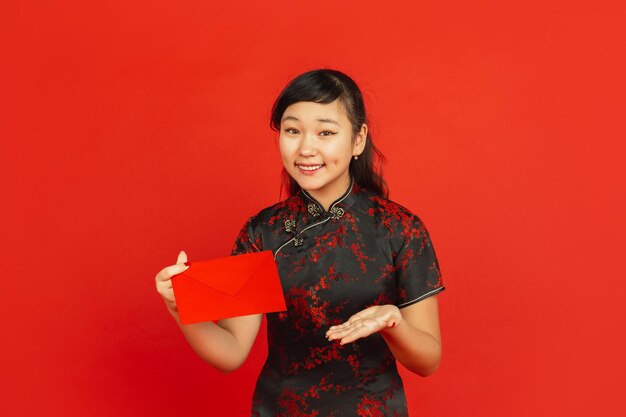 Chinese New Year. Asian young girl's portrait isolated on red background. Female model in traditional clothes looks happy, smiling and showing red envelope. Celebration, holiday, emotions.