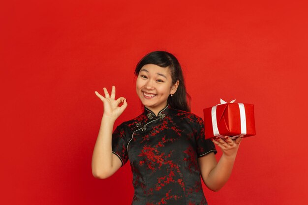 중국의 설날. 빨간색 배경에 고립 된 아시아 젊은 여자의 초상화. 전통적인 옷을 입은 여성 모델은 giftbox와 함께 행복해 보입니다. 축하, 휴일, 감정. 멋지고 웃고 있습니다.