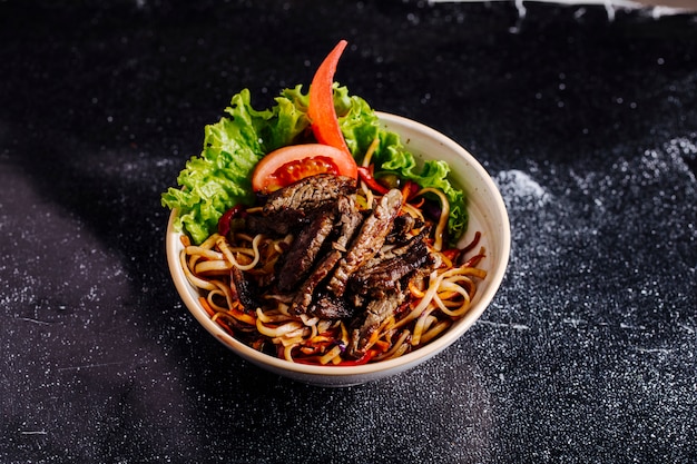 Chinese noodles inside bowl with chopped steak, tomato slices and lettuce.