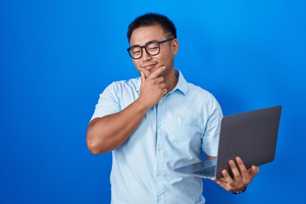 組んだ腕と手をあごに上げてカメラに笑みを浮かべて自信を持って見えるコンピューター ラップトップを使用して中国の若い男。ポジティブ思考。