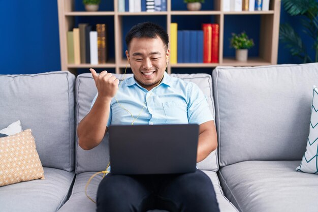 소파에 앉아 컴퓨터 랩탑을 사용하는 중국 젊은이가 입을 벌리고 행복하게 웃는 쪽으로 엄지손가락을 가리키고
