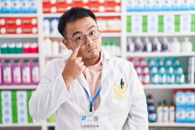 Chinese young man working at pharmacy drugstore pointing to the eye watching you gesture suspicious expression