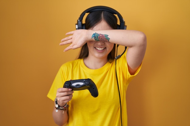 Chinese young woman playing video game holding controller smiling cheerful playing peek a boo with hands showing face. surprised and exited