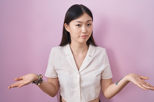 Chinese young woman standing over pink background clueless and confused with open arms, no idea concept.