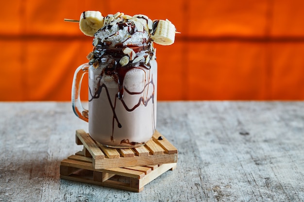 Foto gratuita frullato al cioccolato con sciroppo di cioccolato, banana e panna montata sulla tavola di legno nella superficie luminosa