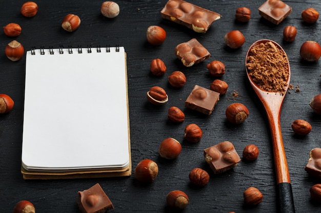 ヘーゼルナッツ​と​チョコレート​、​空白​の​ページ​の​ノート​、​暗い​背景​に​ココア​の​木​の​スプーン​、​シェル​の​ナッツ​に​囲まれて​皮​を​むいた​。