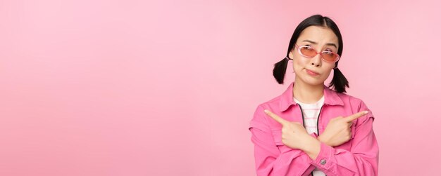 Бесплатное фото Стильная корейская девушка, азиатская модель, указывает пальцами в сторону, показывает два варианта рекламы продукта, демонстрируя предметы, стоящие на розовом фоне
