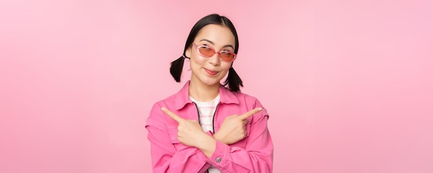 Free photo choice stylish korean girl asian female model points fingers sideways shows two variants product advertisement demonstrating items standing over pink background