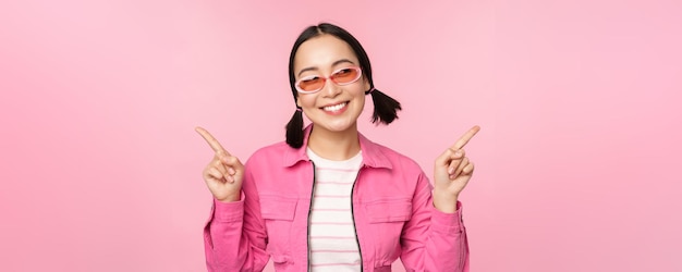 Choice Stylish korean girl asian female model points fingers sideways shows two variants product advertisement demonstrating items standing over pink background