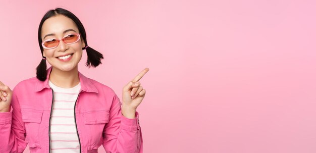 Choice Stylish korean girl asian female model points fingers sideways shows two variants product advertisement demonstrating items standing over pink background