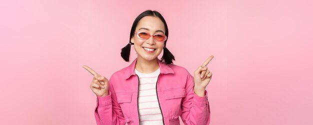 Choice Stylish korean girl asian female model points fingers sideways shows two variants product advertisement demonstrating items standing over pink background