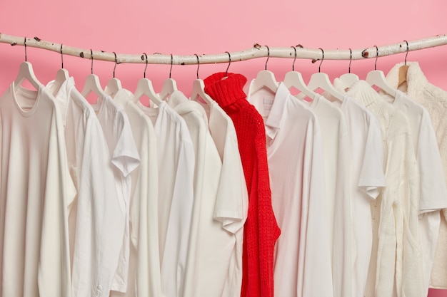 Выбор белой одежды на вешалках в магазине модной одежды. Красный яркий вязаный свитер между нарядами в один тон, изолированный на розовой стене.