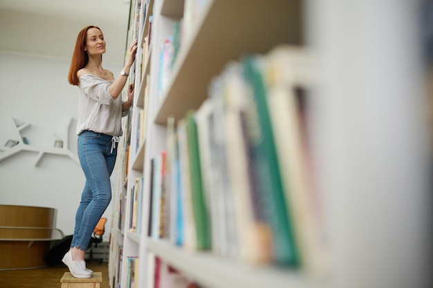 책을 선택합니다. 블라우스와 청바지를 입은 젊고 가느다란 긴 머리 웃는 여성이 도서관에서 책장에 손을 대고 서 있는 책을 찾고 있습니다.