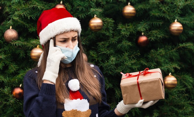 Choosing a gift. christmas mask congratulations. portrait woman wearing santa hat and white sweater in medical mask, giving gift present box with red ribbon, christmas tree bokeh on background