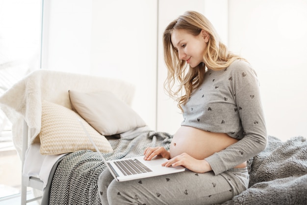 Choosing name for baby. Close up portrait of young attractive blonde pregnant mother in comfy grey home clothes sitting in bedroom, trying to find information about names meaning.