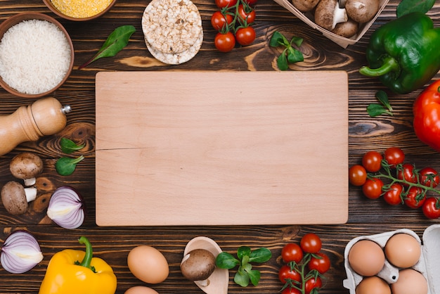 Chopping board surrounded with vegetables; eggs and rice grains on desk