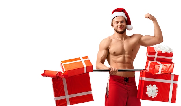 Рождественский бодибилдинг. Обнаженный разорванный парень в шляпе Санта-Клауса держит штангу с подарками, демонстрируя свои мышцы в позе бодибилдинга, изолированной на белом. 2018, 2019.
