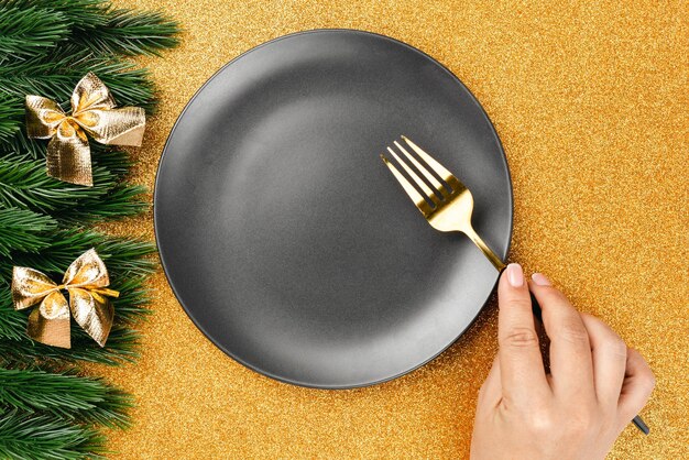 Christmas card flat lay woman hand holding fork on black empty plate on golden background with frame...