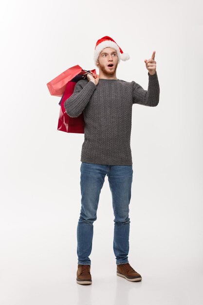 Концепция Рождества привлекательный молодой кавказский мужчина удивительно шокирует, держа сумку с покупками и указывая пальцем впереди
