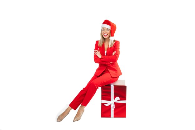Christmas concept photo of elegant blonde lady in red suit and high heels sitting on wrapped gift box. Winter holiday concept