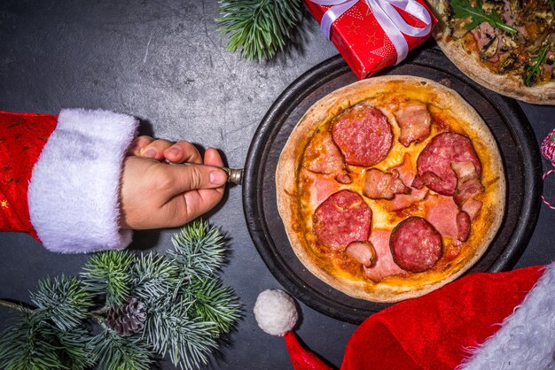ペパロニとキノコのピザでクリスマスに飾られた背景、サンタの手でピザのスライスを取ります。配達、レストランのクリスマスランチパーティーメニュー、サンタはクリスマスにピザを配達し、上の黒いテーブル Premium写真