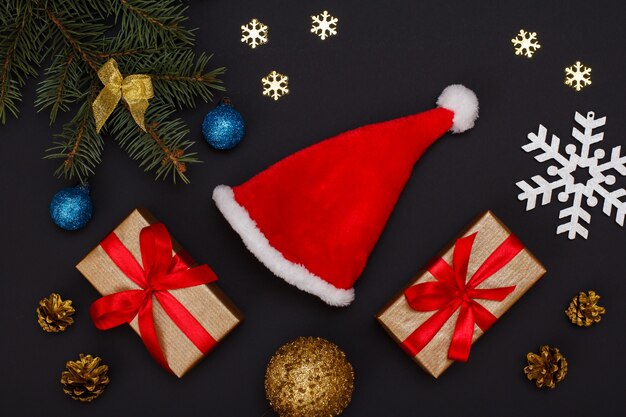 크리스마스 장식. 산타 모자, 선물 상자, 원뿔이 있는 전나무 나뭇가지와 검은 배경에 장식용 눈송이가 있습니다. 평면도. 크리스마스 인사말 카드 개념입니다.
