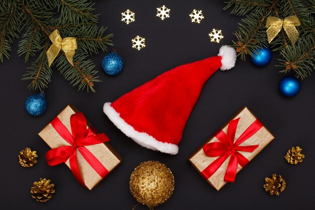크리스마스 장식. 산타 모자, 선물 상자, 원뿔이 있는 전나무 나뭇가지와 검은 배경에 장식용 눈송이가 있습니다. 평면도. 크리스마스 인사말 카드 개념입니다.