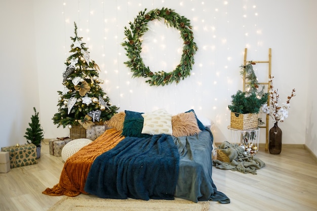 회색과 흰색 색상의 크리스마스 축제 인테리어입니다. 둥근 녹색 화환이 벽에 걸려 있습니다. 포근한 담요로 덮인 침대가 화환으로 장식된 집에 서 있다.