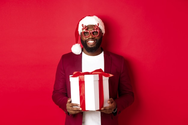 Natale. bell'uomo afroamericano in occhiali da festa e cappello da babbo natale con regalo di capodanno, porta un regalo in scatola e sorridente, in piedi su sfondo rosso