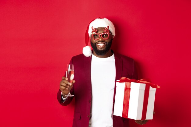 クリスマス。パーティーグラスとサンタ帽子、新年の贈り物とシャンパングラスを持って、幸せな休日、赤い背景を願ってハンサムなアフリカ系アメリカ人の男