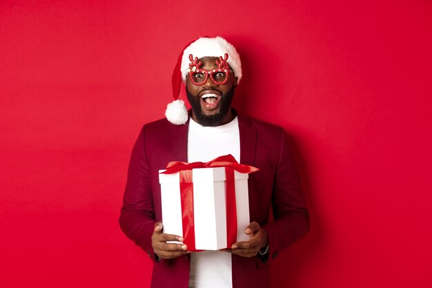 Christmas. Handsome african american man in party glasses and santa hat holding new year gift