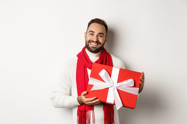 Christmas. Handsome young man holding gift and smiling, wishing happy holidays and giving presents, standing over white background
