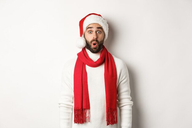 Christmas holidays. Bearded man looking surprised at camera, wearing santa party hat and red scarf, standing against white background
