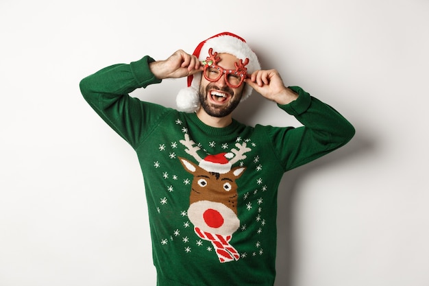 Бесплатное фото Рождественские праздники, концепция празднования. счастливый человек в шляпе санта-клауса и забавных партийных очках, стоящих на белом фоне