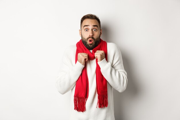 クリスマス休暇とお祝いのコンセプト。カメラに畏敬の念を抱き、驚いて立って、新年に寒さを感じ、赤いスカーフとセーター、白い背景を身に着けている男