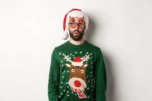 Christmas holidays, celebration concept. surprised guy in party glasses and santa hat looking down amused, standing over white background